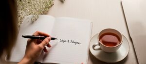 Uma cena criativa sobre a mesa mostra uma mão segurando uma caneta e escrevendo em um caderno as palavras "Logo e Slogan"